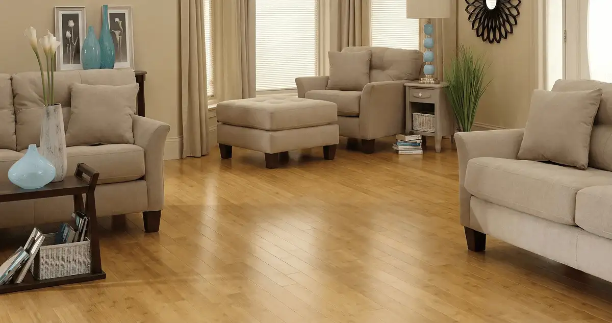 A living room with professionally installed bamboo flooring, beige sofas, a matching ottoman, a side table with a blue lamp, vases, framed pictures, and large windows with blinds and curtains in Fargo, ND